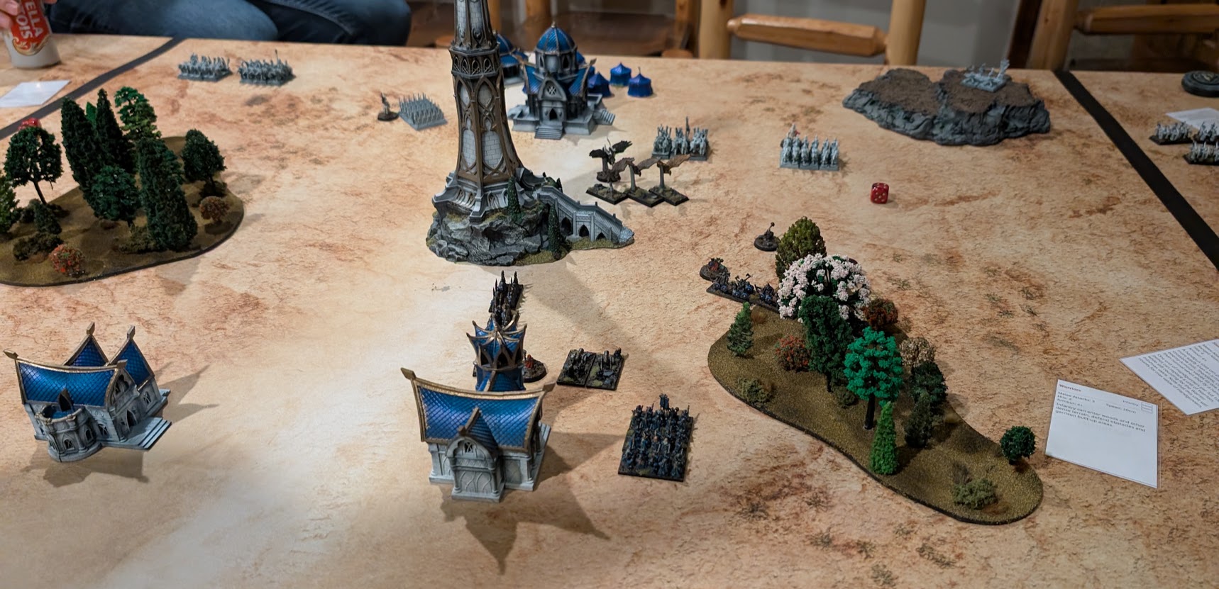 Warmaster units maneuver among blue-roofed elven buildings, a tall tower, and patches of woodland.